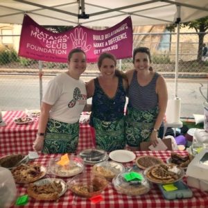 3 women with aprons smile with arms around each other behind a table laden with pies. A sign behind them reads Batavia Mothers' Club Foundation, at the heart of the community. "Build relationships, Make a Difference, Get Involved"